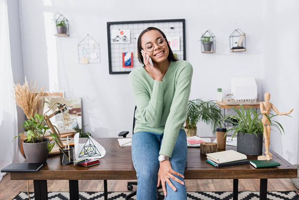 boldog afro-amerikai építész beszél okostelefonon, miközben ül az asztalon otthon stúdió - Fotó, kép