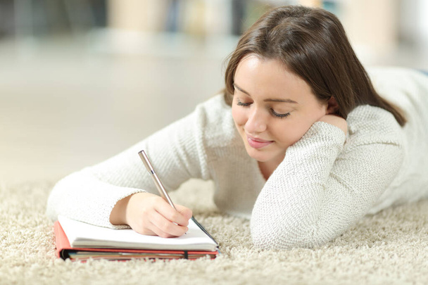 Happy teen writing notes on notebook lying on a carpet on the floor at home - Fotografie, Obrázek
