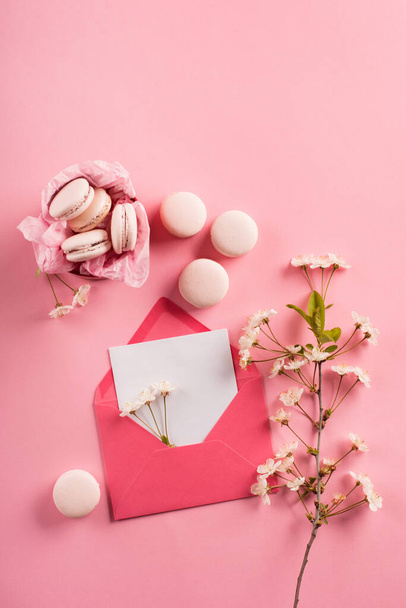Festive holiday greeting card for Valentines, Birthday, Woman, Mothers Day. Rose macaroons in gift box, Cherry blossoms, empty envelope. Sweet macarons present on pink background. Top view, copy space - Foto, imagen