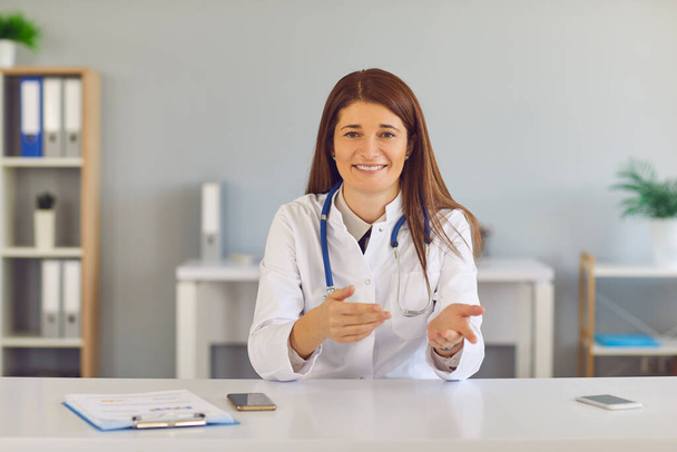 Smiling young woman doctor sitting and gesticulating during online meeting with patient - 写真・画像