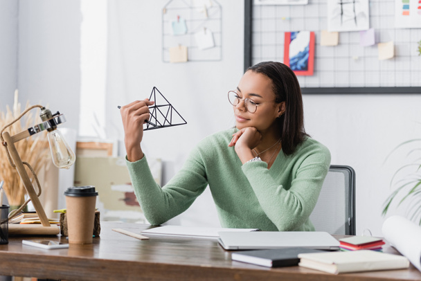 african american architect in eyeglasses holding model of pyramid near coffee to go and notebooks on blurred foreground - Photo, Image
