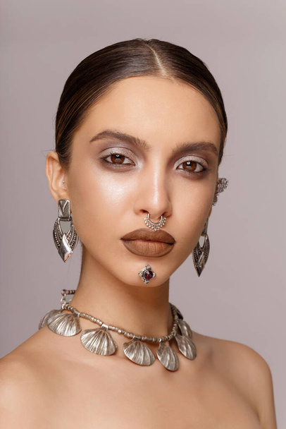 Frontaal portret van een meisje met make-up en sieraden aangebracht op het gezicht, met blote schouders, over grijze achtergrond. - Foto, afbeelding