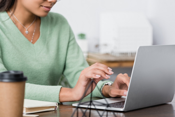 bijgesneden weergave van Afrikaanse Amerikaanse interieur ontwerper werken op laptop in de buurt van koffie te gaan op wazig voorgrond - Foto, afbeelding