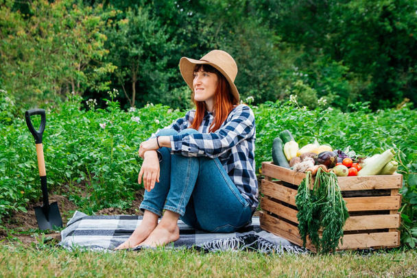 Hermosa agricultora sentada en una manta cerca de verduras orgánicas frescas en una caja de madera sobre el fondo de un huerto. - Foto, imagen