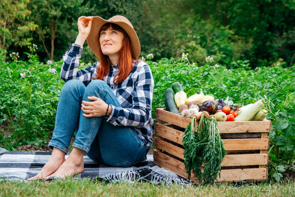 Beautiful female farmer sitting on a blanket near fresh organic vegetables in a wooden box against the background of a vegetable garden. - Photo, Image