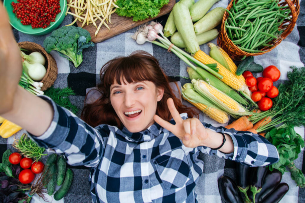 Portrait beautiful woman taking self-portrait lying on a plaid among fresh organic vegetables. Top view. Funny face. - Photo, Image