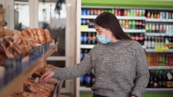 Una donna che indossa una maschera protettiva medica compra cibo in un supermercato o negozio di alimentari. Il cliente mette il pane in un cestino - Filmati, video
