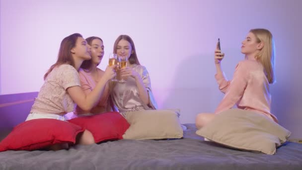 Cuatro hermosas mujeres jóvenes felices en pijama se sienta en la cama, bebe vino y toma fotos en la despedida de soltera - Metraje, vídeo