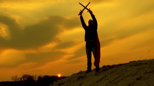 Game of Roman legion. Free male knight crossed his swords in sky above his head. Superman with a sword in his hand and in a red cloak stands on mountain in sunset light. Free man plays a superhero. - Footage, Video