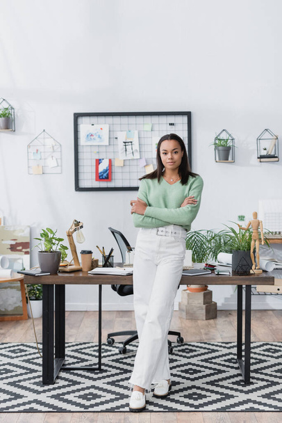young african american architect looking at camera while standing near desk with crossed arms - Valokuva, kuva