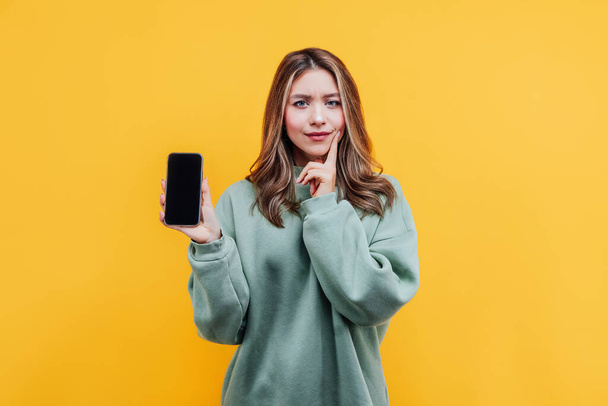 Pretty girl on a yellow background holds a phone with a black screen and looks thoughtfully at the camera. Studio photo. Commercial offer. Place for text. - Фото, изображение