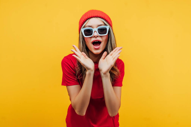 Modern girl in glasses and a red hat on a yellow background, makes a surprised face, wearing sunglasses. Place for text. Commercial offer. - Photo, image