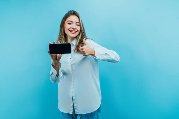 Photo of an attractive young girl holding a phone with a black screen in her hand and showing the class smiling at the camera Blue background, studio photo. Commerce, Photo for advertising. - Φωτογραφία, εικόνα