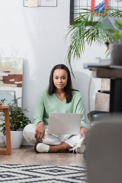 young african american interior designer looking at camera while sitting on floor with laptop, blurred foreground - Photo, Image