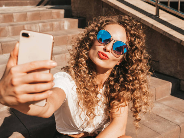 Beautiful smiling model with afro curls hairstyle dressed in summer hipster clothes.Sexy carefree model sitting on  stairs in the street in sunglasses.Taking selfie self portrait photos on smartphone - Photo, Image
