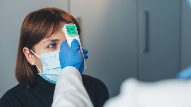 Il medico caucasico che indossa i guanti sta misurando la temperatura corporea del paziente con maschera durante una consultazione. - Foto, immagini