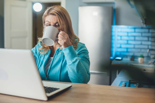 Busy senior blonde woman is drinking a cup of tea while working from home at the laptop in the kitchen - Photo, Image