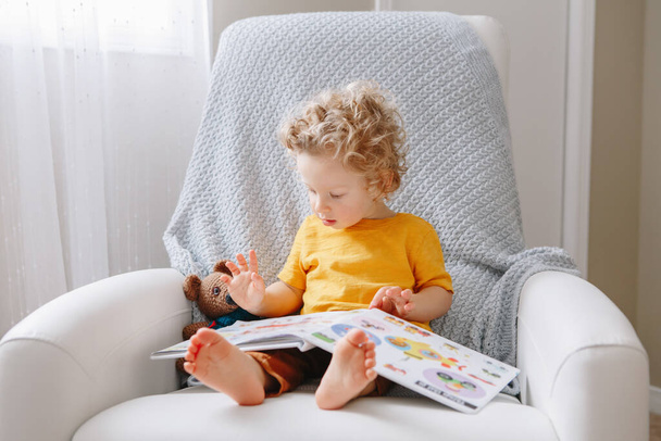 Cute adorable little baby boy toddler 2 years old sitting in armchair at kids room and reading book. Early age kid child development education. Candid home authentic childhood lifestyle - Photo, Image