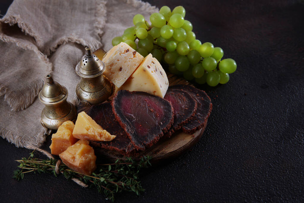 Homemade sausage and cold cuts on a black background. Craft cheese and green grapes on a wooden board. Natural products. Wooden boards for serving food. Top view. Copy space. - Foto, afbeelding