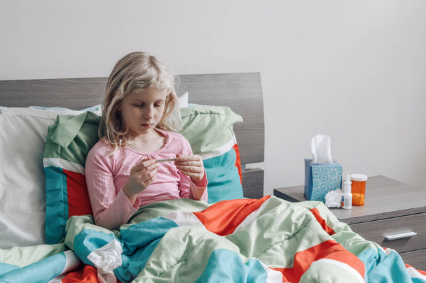 Sick ill Caucasian girl with fever lying in bed at home blowing nose with paper tissue and checking temperature on thermometer. Virus cold season flu illness. Medicine and health concept - Fotografie, Obrázek