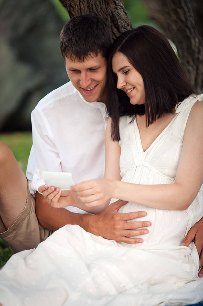 Una ragazza incinta e un ragazzo siedono sotto un albero nel parco e guardano la foto ad ultrasuoni del nascituro..  - Foto, immagini