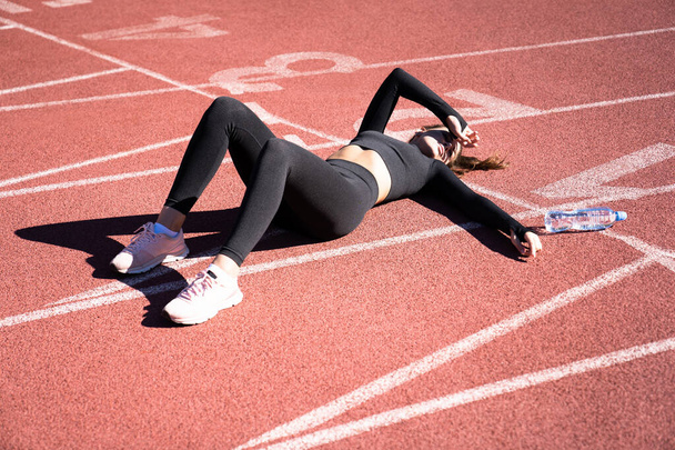 Top view of tired fit woman in sportswear resting after workout or running on a treadmill rubber stadium, holding a bottle of water, taking a break during training, outdoors. - Fotoğraf, Görsel