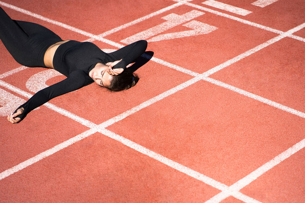 Верхний вид уставшей женщины в спортивной одежде отдыхающей после тренировки или бега на резиновом стадионе беговой дорожки, держащей бутылку воды, отдыхающей во время тренировки, на открытом воздухе. копировальное пространство - Фото, изображение