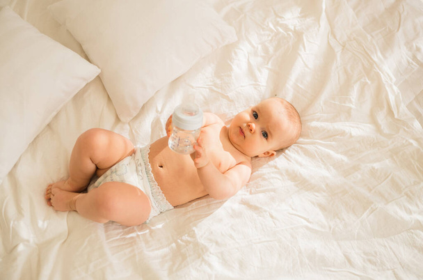 Bir kız bebek bezi içinde beyaz yatak çarşaflarının üzerinde bir şişe suyla yatıyor. - Fotoğraf, Görsel