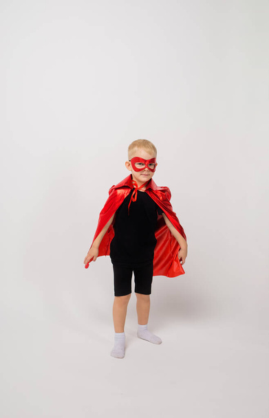 un niño pequeño en un disfraz de superhéroe se para sobre un fondo blanco y mira a la cámara con un lugar para el texto - Foto, Imagen