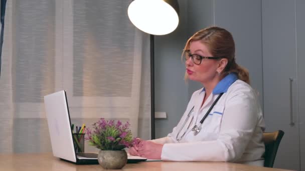 Sivukuva video valkoihoisesta lääkäristä lääketieteellisessä takissa puhumassa kannettavalla potilaan kanssa selittäen parannusvaiheet - Materiaali, video