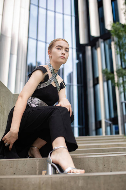 European young woman sitting on concrete stairs near metal panels - 写真・画像