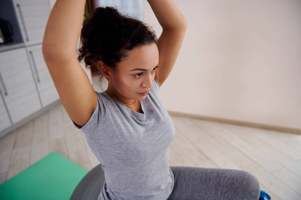 Mujer deportiva sentada en una pelota de fitness y levantando los brazos mientras hace ejercicio - Foto, imagen