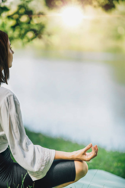 Młoda kobieta siedząca w pozycji lotosu i praktykująca medytację w pobliżu wody w przyrodzie - Zdjęcie, obraz