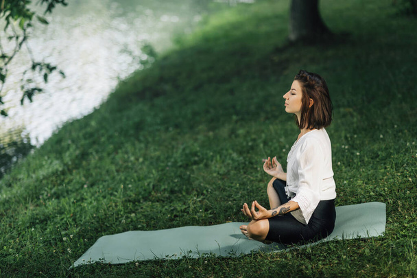 Młoda kobieta siedząca w pozycji lotosu i praktykująca medytację w pobliżu wody w przyrodzie - Zdjęcie, obraz