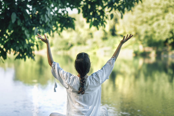 Itsetutkiskelua luonnossa järven rannalla. Lootuksessa istuva nainen meditoi avosylin.  - Valokuva, kuva