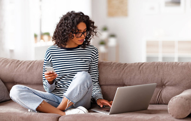 Etnikai női böngészés okostelefon és laptop, miközben ül keresztbe lábú kényelmes kanapén otthon - Fotó, kép