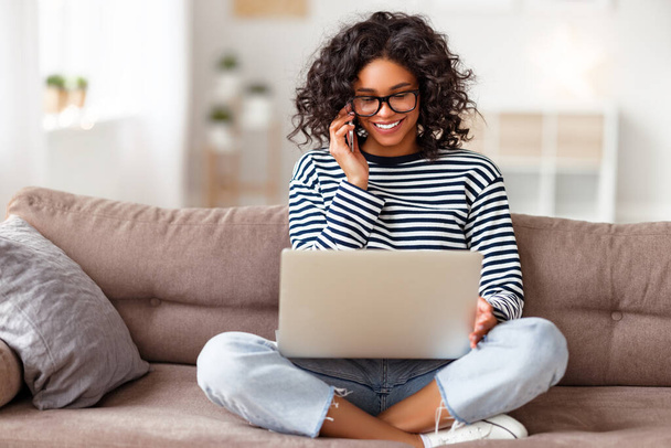 Onnellinen etninen nainen käyttää kannettavaa tietokonetta ja puhuu älypuhelimella istuessaan sohvalla viihtyisässä olohuoneessa kotona - Valokuva, kuva