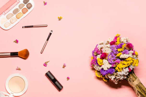 Επίπεδη φωτογραφία lay με πολύχρωμο μπουκέτο λουλουδιών και μακιγιάζ: λαμπερή σκιά ματιών, πινέλα, κραγιόν και πούδρα σε ροζ φόντο. - Φωτογραφία, εικόνα