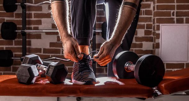 Fitness motivation and muscle training concept. Man in sneakers tying shoelaces in sunlight. Athlete starting exercise with dubbell weight. - Foto, immagini
