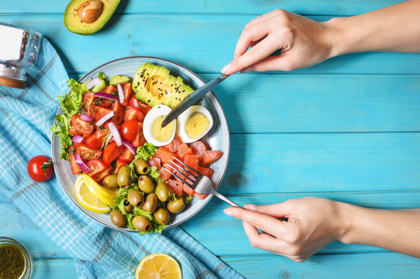 Ketogenic, paleo diet lunch bowl with salted salmon fish, lemon, avocado, olives, boiled egg, tomato, green lettuce salad. Female hands holds fork and knife. top view - Photo, Image
