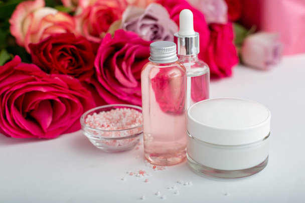 Glass cosmetic bottles, cream, serum, soap, oil on white table floral background. Flower red pink roses natural organic beauty product. Spa, skin care, bath body treatment. Set of cosmetics with rose - Fotografie, Obrázek