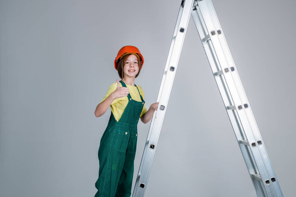 cheerful teen girl in working uniform and helmet on ladder show thumb up, building - Фото, зображення