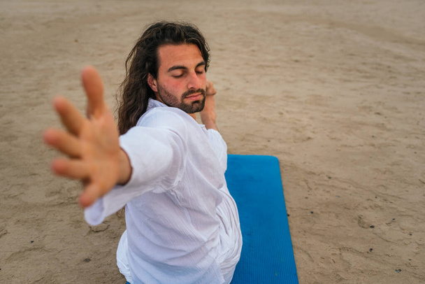 Uzun saçlı yetişkin bir adamın plajda yoga pozu verirken çekilmiş resmi.. - Fotoğraf, Görsel