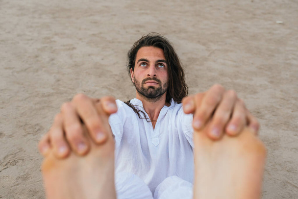 Uzun saçlı yetişkin bir adamın plajda yoga pozu verirken çekilmiş resmi.. - Fotoğraf, Görsel