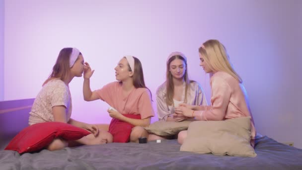 Neljä onnellista kaunista nuorta naista yöpuvussa ja otsanauhat päällä istuu sängyllä ja soveltaa Eye Collagen laastaria ja kermaa polttareissa - Materiaali, video