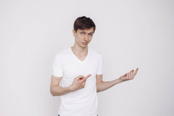 Портрет молодого человека в белой футболке, который показывает что-то, изолированное на белом фоне. - Фото, изображение