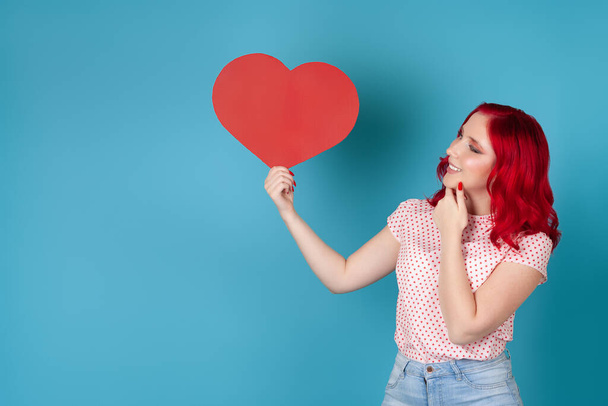 a dreamy contented young woman with red hair holds a red paper heart and scratches her chin with her hand , isolated on a blue background - Фото, изображение