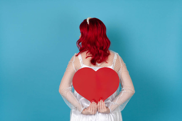 una joven con un vestido blanco y pelo rojo sostiene un gran corazón de papel rojo detrás de su espalda ocultando su cara, aislada sobre un fondo azul - Foto, imagen