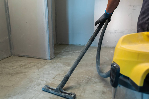 Ο κατασκευαστής καθαρίζει το πάτωμα από τη σκόνη με μια ηλεκτρική σκούπα κατασκευής. - Φωτογραφία, εικόνα