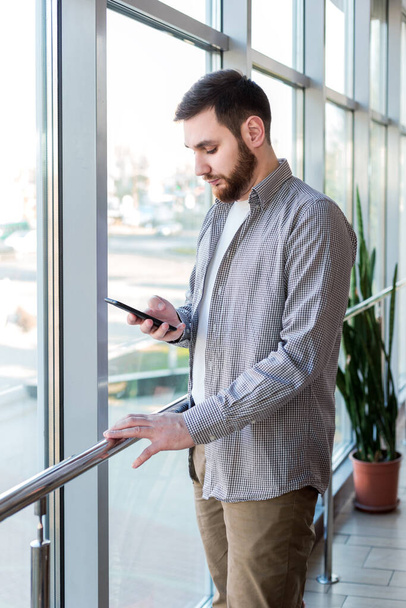 Smart beard Caucasian man is chatting texting on smartphone in urban modern coworking office on glass balcony near panoramic window. - Zdjęcie, obraz